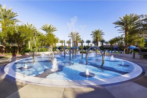 Una fuente circular con múltiples chorros, rodeada de palmeras y un cielo despejado.