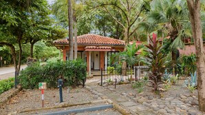 A small, single-story building with a tiled roof, surrounded by lush greenery and a gravel pathway. - Studio across the street from beach, top location (Playa Langosta)