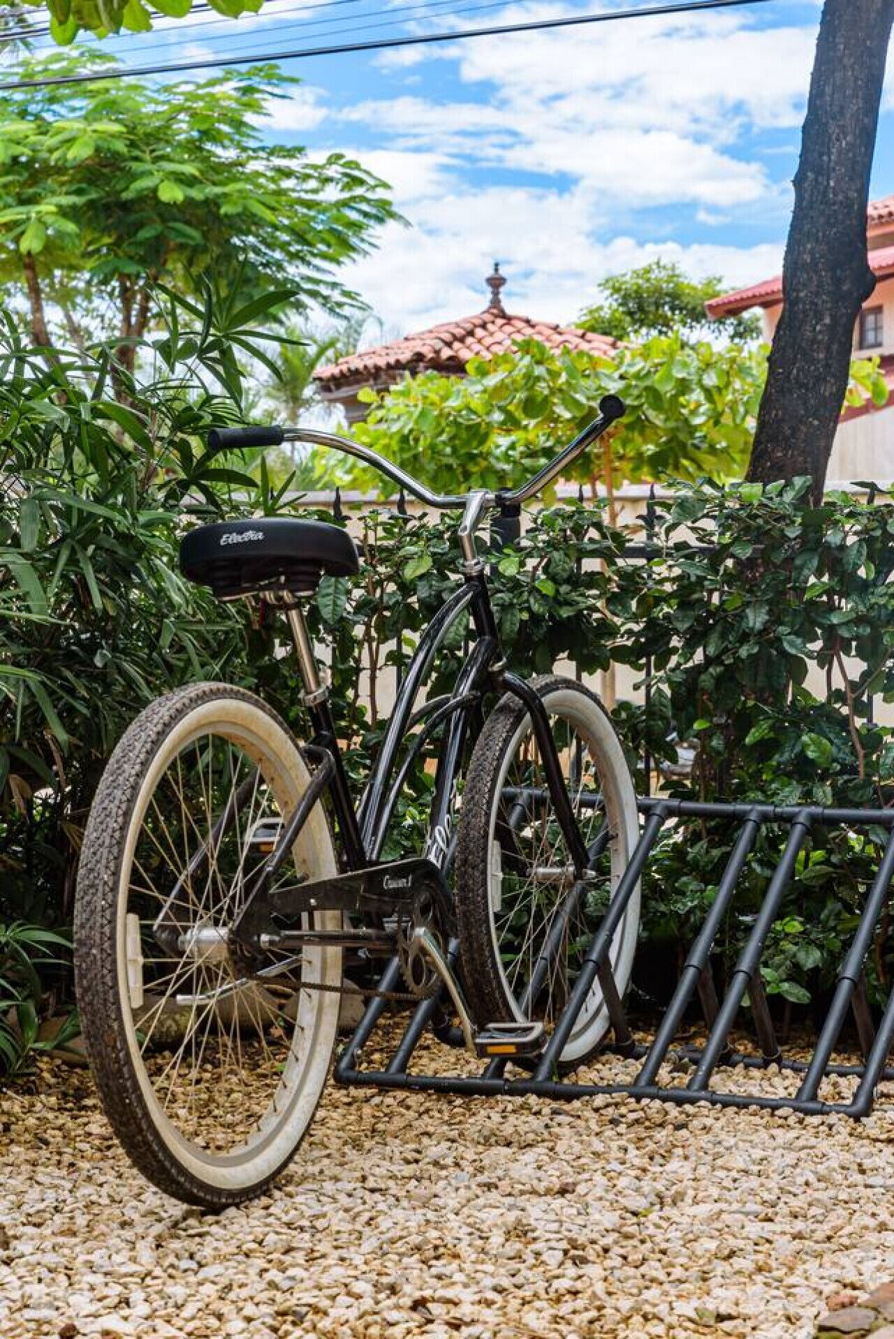 Una bicicleta asegurada a un soporte, con suelo de grava y follaje al fondo.