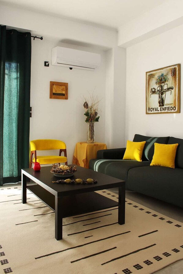 A living room with a black coffee table, a yellow armchair, a dark sofa with yellow cushions, a decorative vase, and a framed picture on the wall.