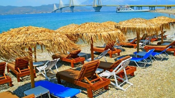 A beach with thatched umbrellas and wooden lounge chairs. A bridge is visible in the background. - Central Old Town Stay - Bright & Fully Equipped (Patras)