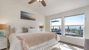 A bedroom with a bed, a ceiling fan, a painting, two bedside tables, and a view of the sea through large windows. - Bodega Bay Oasis  - Wine Tastings Included (Bodega Bay)