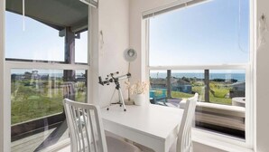 Una habitación con un escritorio blanco, dos sillas blancas, un telescopio sobre un trípode, una planta en maceta y ventanas amplias que dan a vistas del mar y edificios.
