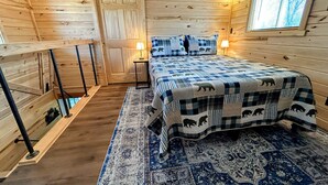 A cozy cabin-style bedroom with a large bed, a wooden door, a window, and a small table with a lamp.