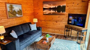 A cozy living room with a dark sofa, two side tables with lamps, a coffee table with a plant, a large TV, and a painting of a forest on the wall.