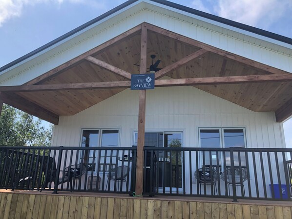 Une charmante maison de campagne située en bord de mer, appelée « The Bayview », dotée d’une terrasse en bois, d’une rambarde noire et d’un patio couvert.
