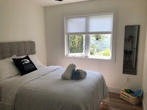 Une chambre à coucher avec un lit, une fenêtre avec des stores, une tête de lit, un oreiller orné d’un motif, une petite table avec des bouteilles et une caisse en bois contenant des jouets.