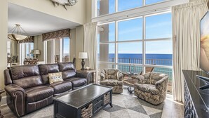 A spacious living room with a large window overlooking the ocean, a leather sofa, patterned armchairs, and a flat-screen TV.