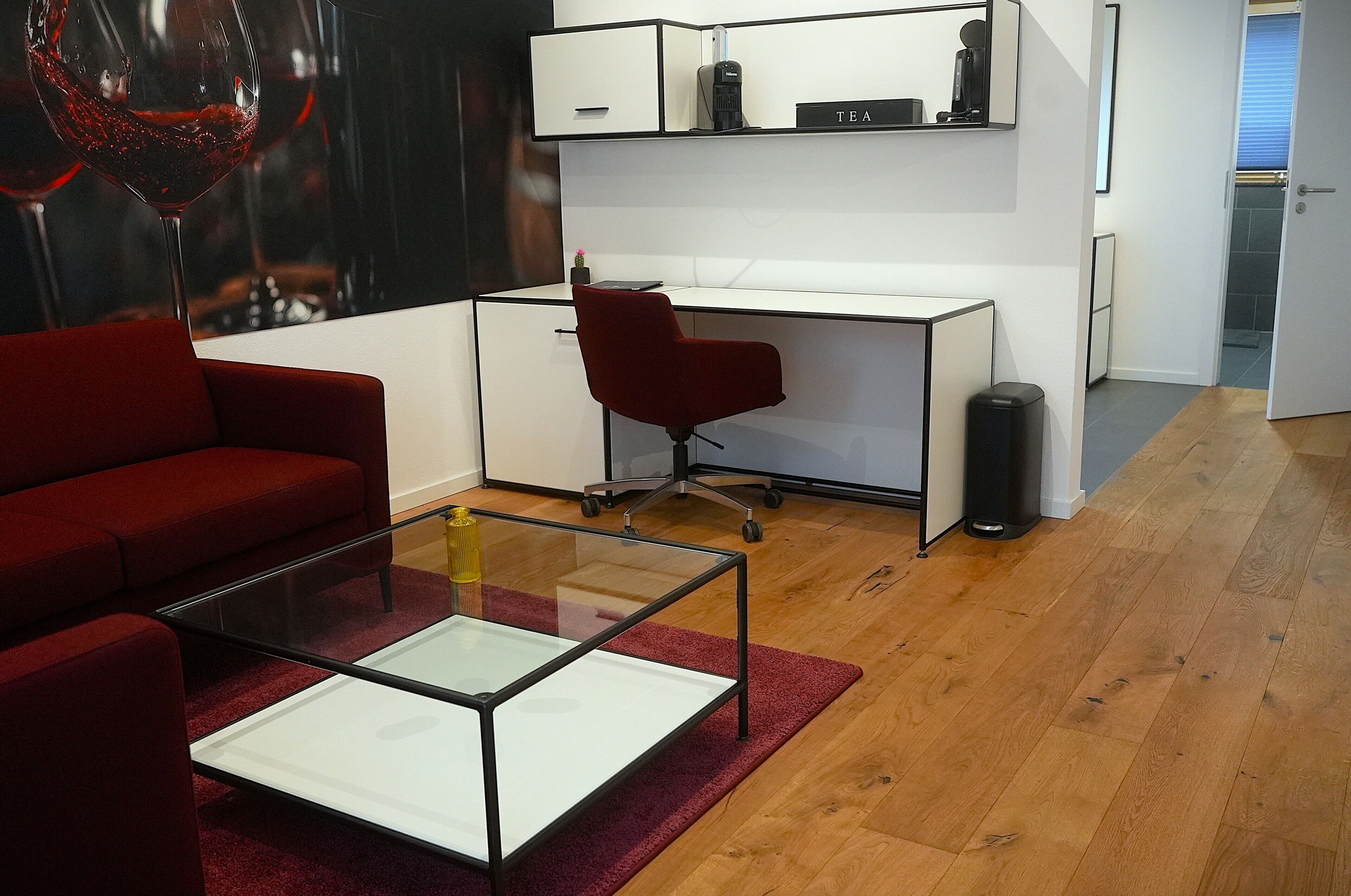 Una sala de estar moderna con un sofá rojo, una mesa de centro de vidrio, una silla de oficina roja, un escritorio blanco y un bote de basura.
