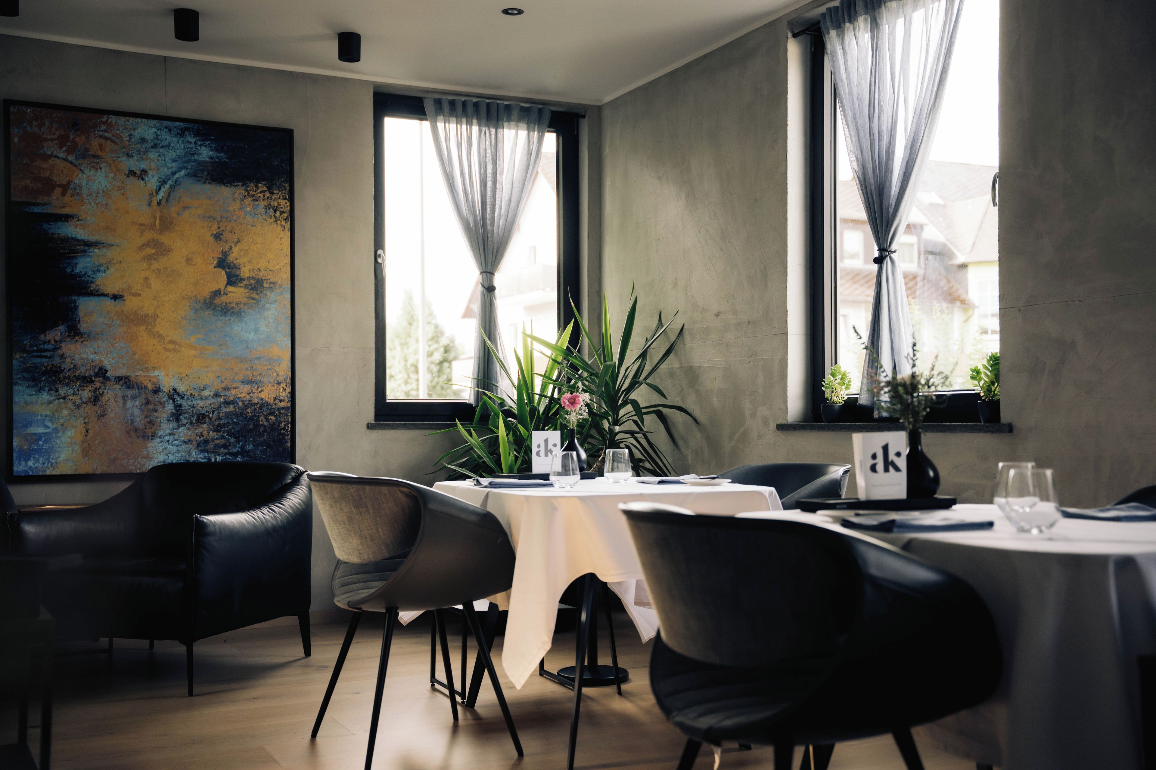 Een moderne eetruimte met een tafel gedekt voor twee personen, omringd door stoelen, met een groot abstract schilderij aan de muur en uitzicht op het exterieur door grote ramen.