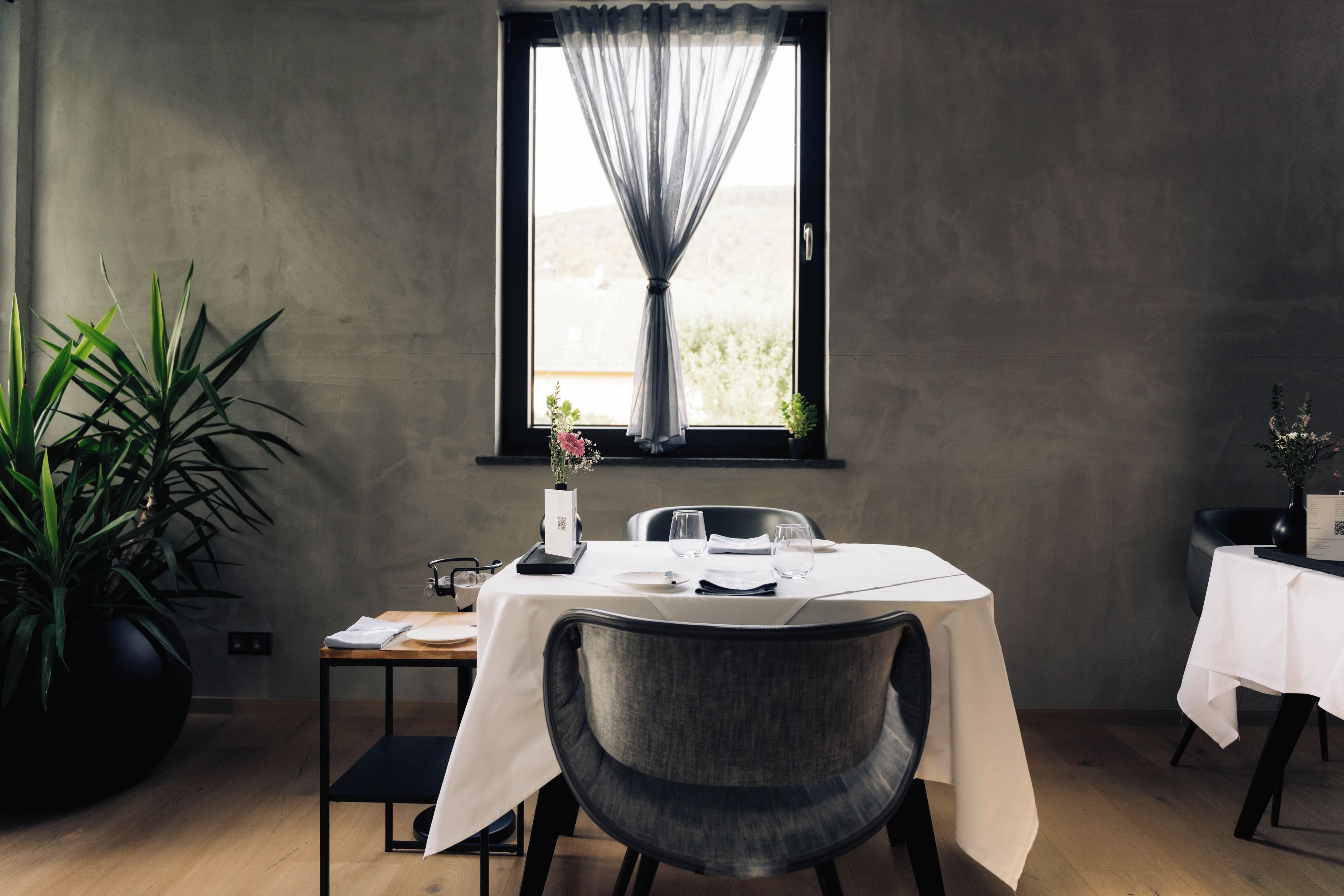 A dining area with a table set for two, a potted plant, and a window with sheer curtains.