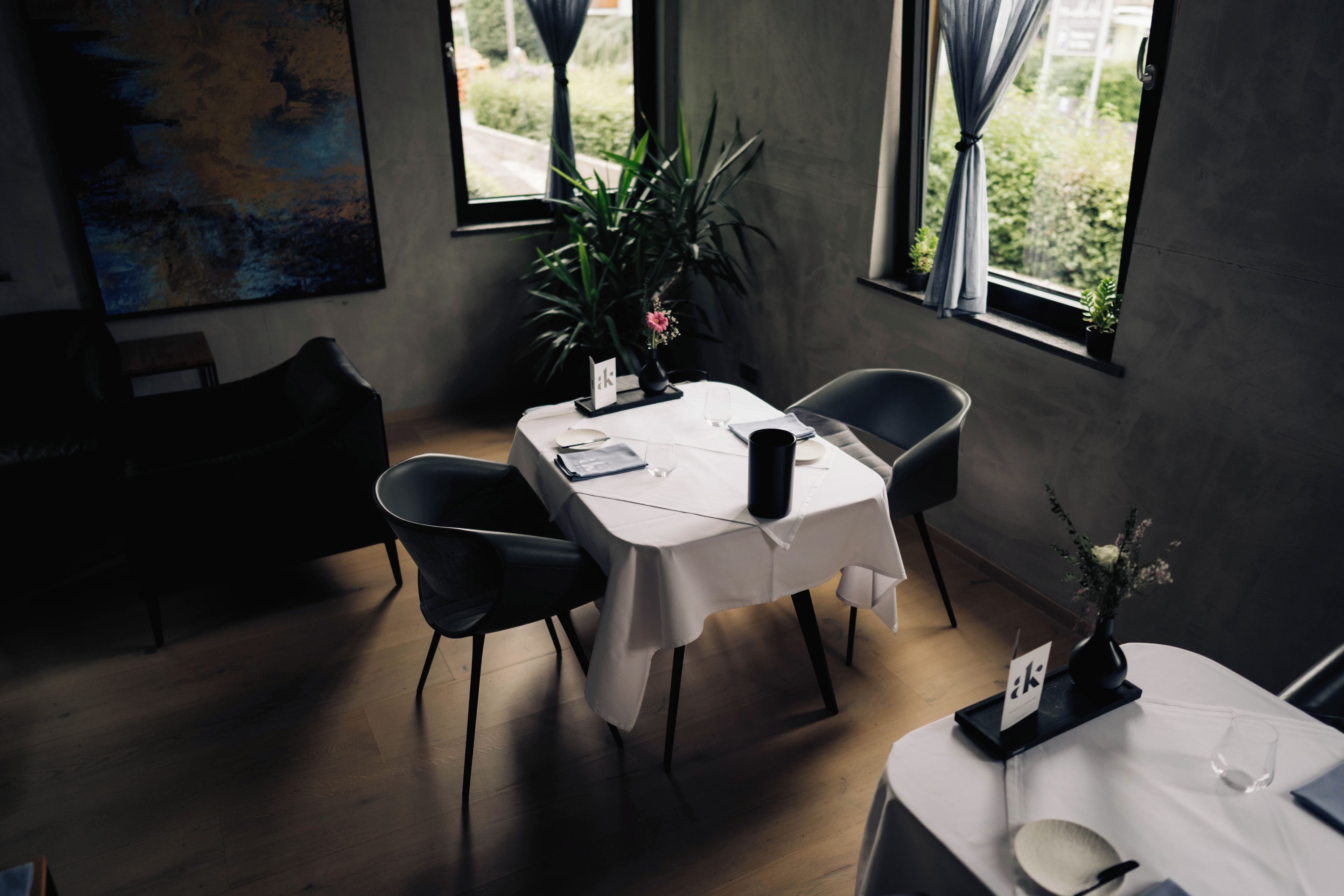 Un coin repas avec une table dressée pour deux personnes, des chaises, une plante en pot et une fenêtre avec des rideaux.