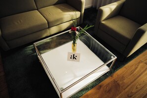 Una mesa de centro con superficie de vidrio, un logo, un jarrón con flores y un piso de madera.