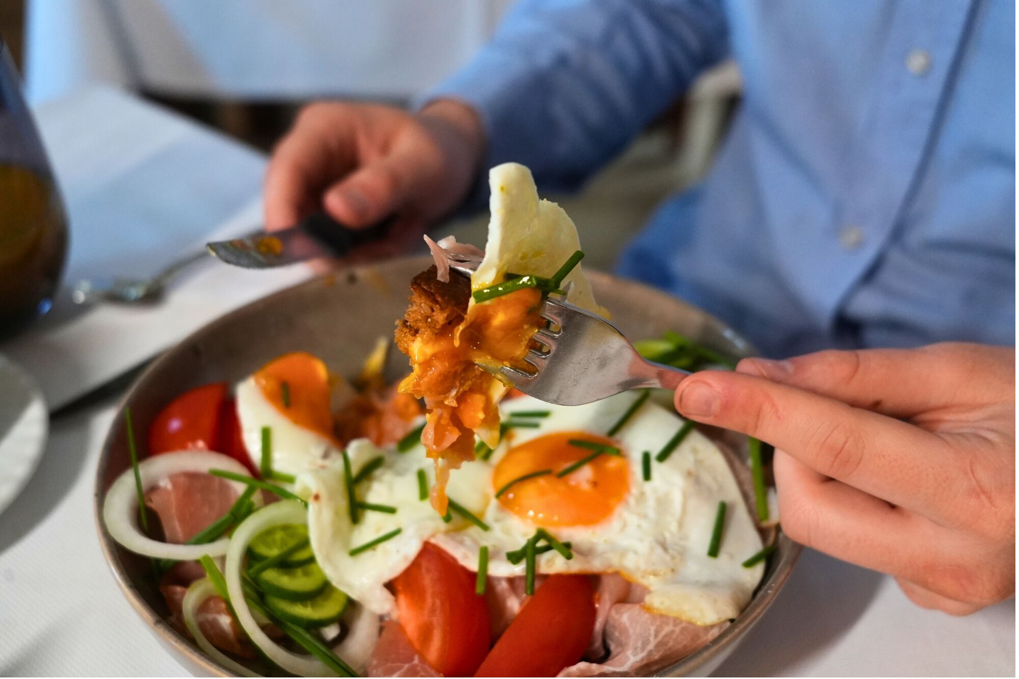 Une personne tenant une fourchette avec un morceau de nourriture, peut-être un sandwich ou un morceau de viande, au-dessus d’une assiette contenant des œufs, des légumes et une sauce.