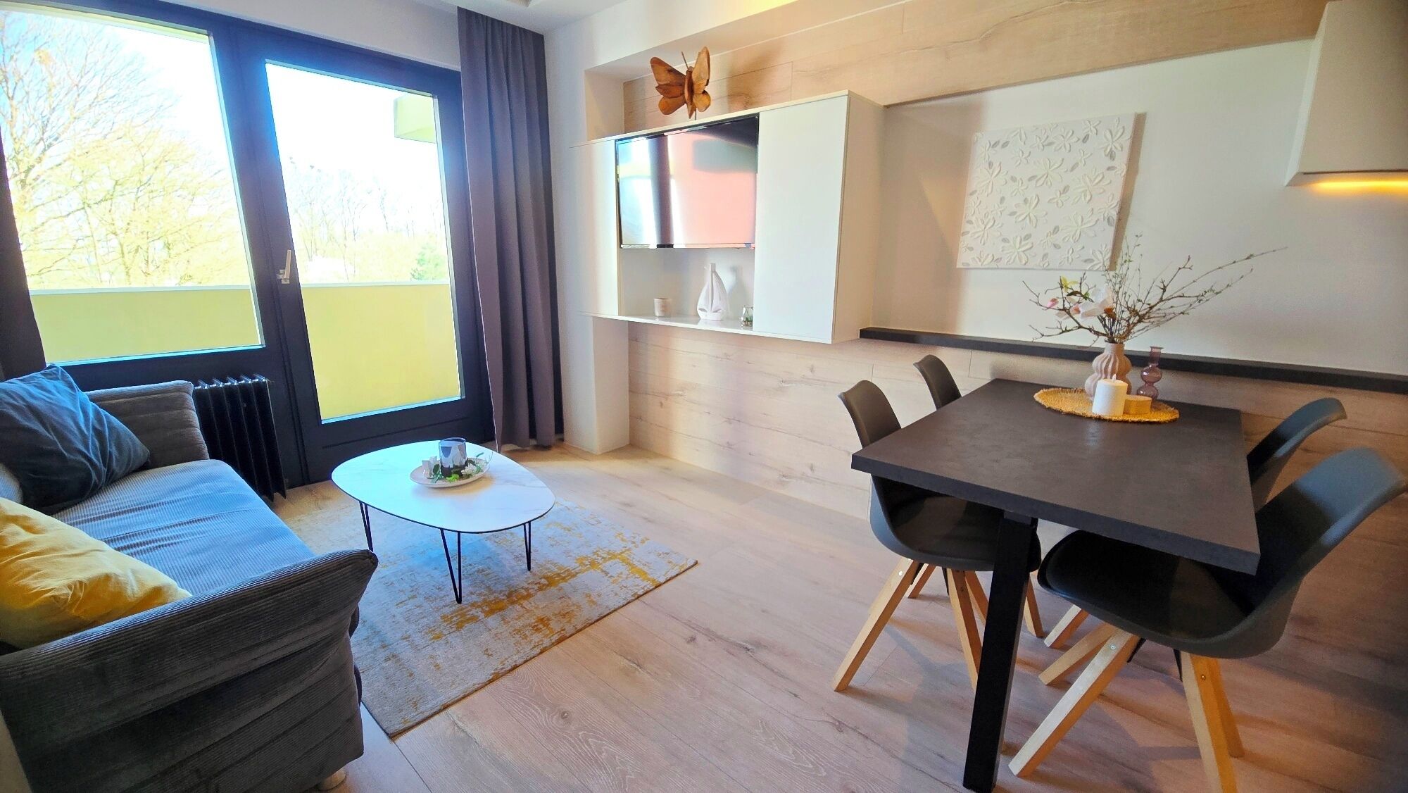 A modern living room with a sofa, coffee table, and dining table. There is a window with curtains, a wall-mounted shelf with decorative items, and a view of trees outside.