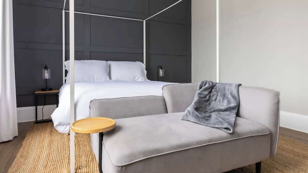 A modern bedroom with a four-poster bed, a grey sofa, a small wooden side table, and a dark wall paneling. - Hotel Quality/ Walkable / Free Parking/ Office #11 (Lakewood)