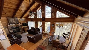 A cozy cabin interior with a stone fireplace, a dining area, and a living room with a sofa and chairs.