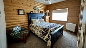 A wooden-paneled bedroom with a bed, a chair, a nightstand, a lamp, a window, and a ceiling fan.
