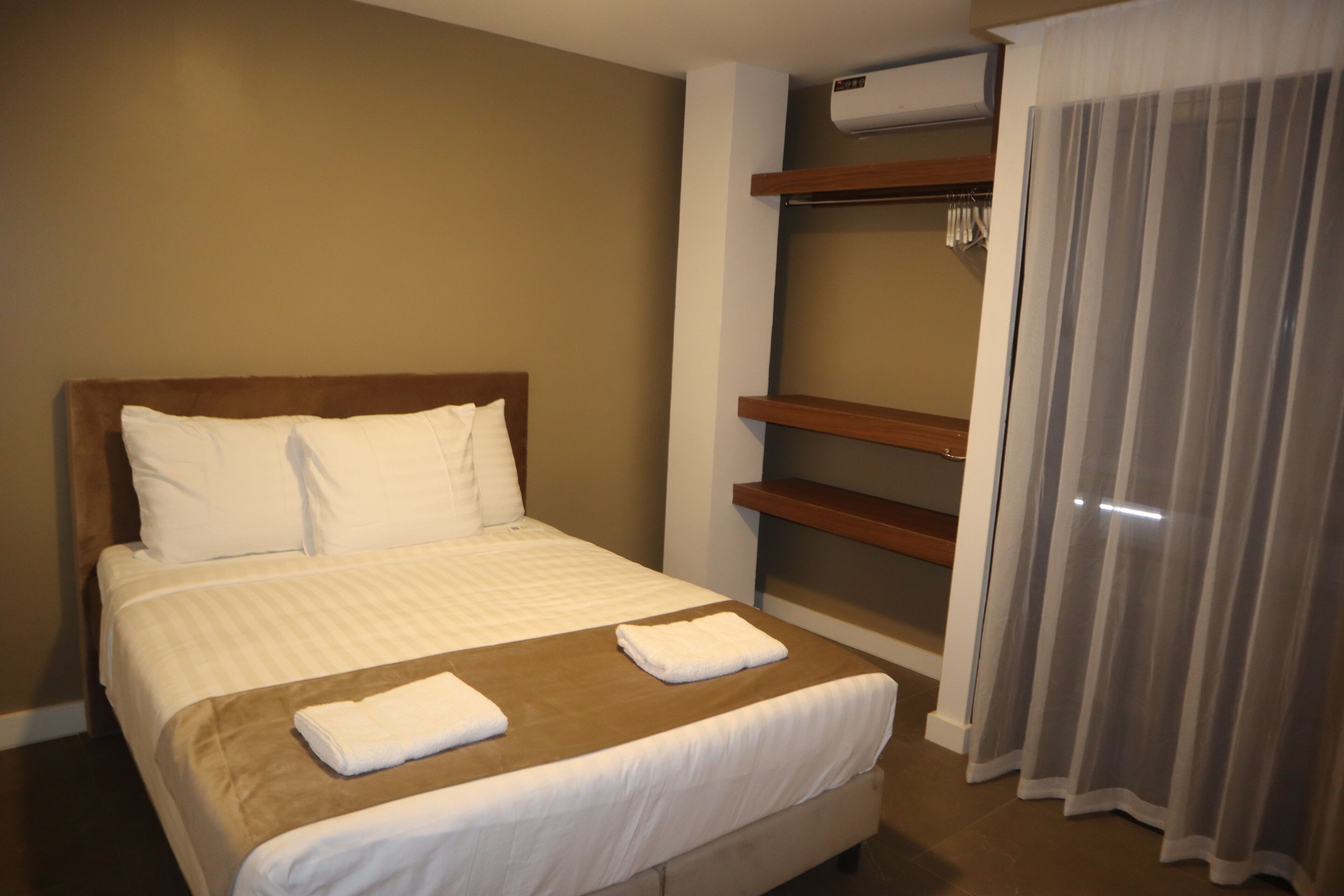 Une chambre d’hôtel avec un lit, une tête de lit en bois, une étagère intégrée et une fenêtre avec des rideaux.