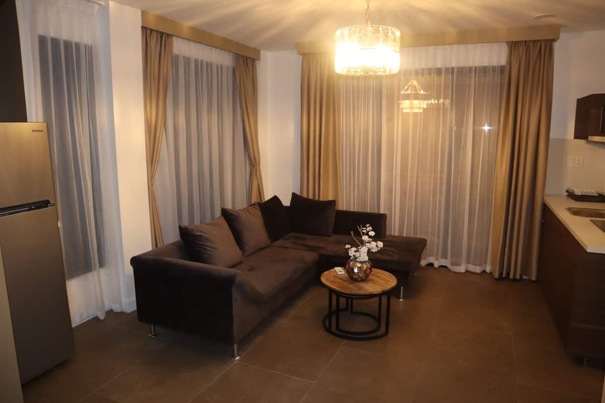 A modern living room with a dark brown sofa, a small round coffee table with a vase, and a kitchen area with a refrigerator and counter.