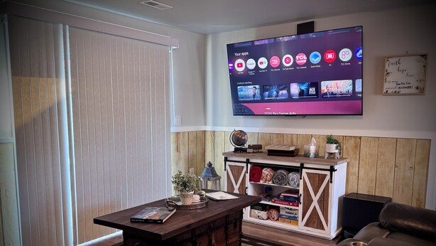 Ein Wohnzimmer mit an der Wand montiertem Fernseher, einem Holzschrank mit diversen Gegenständen, einem Ledersofa und einem gemusterten Teppich.