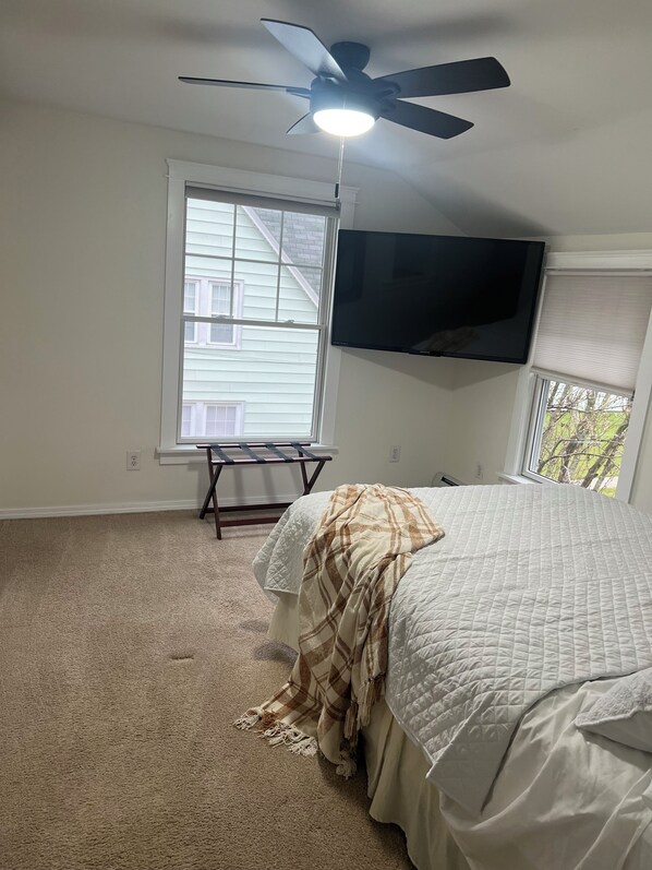 A bedroom with a bed, a ceiling fan, a TV mounted on the wall, and a window with blinds.