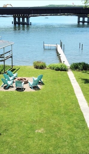 A grassy area with a dock extending into a body of water, a bridge in the background, and a gazebo with chairs.