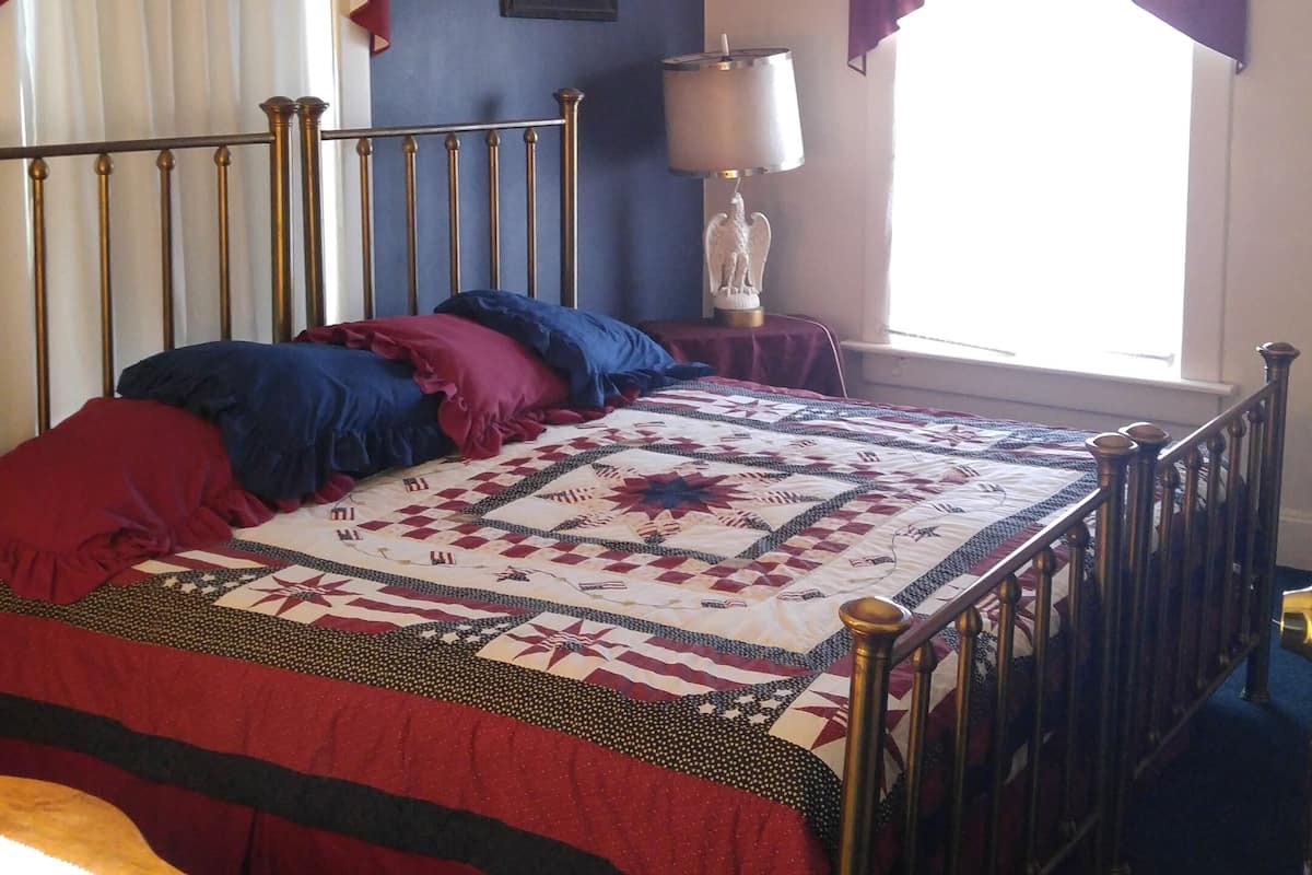A bedroom with a bed, a nightstand, a lamp, a framed picture, and a window with curtains.