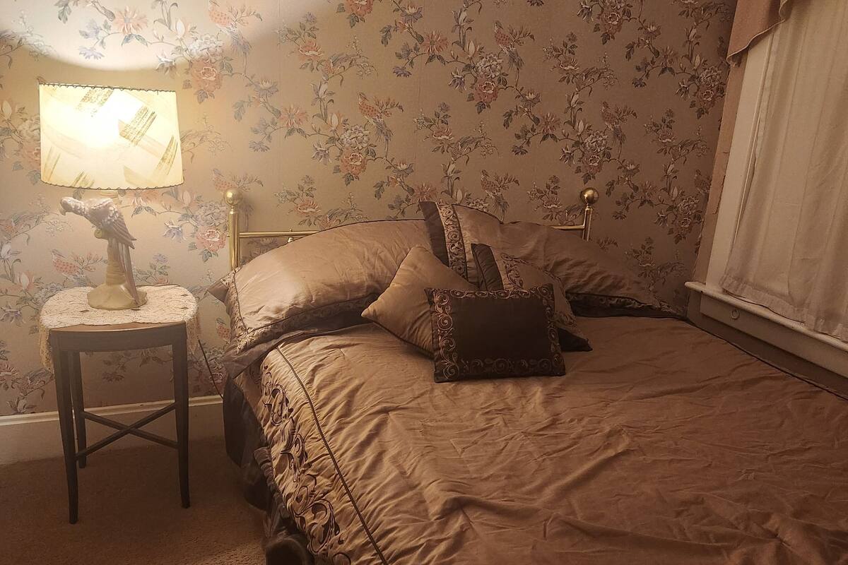 A bedroom with floral wallpaper, a bed with a beige bedspread, a nightstand with a lamp, and a window with curtains.