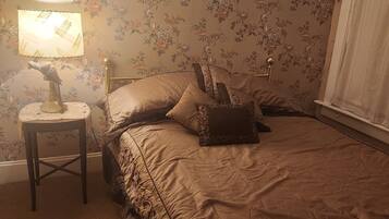 Un dormitorio con papel tapiz floral, una cama con cubrecama color beige, una mesita de noche con una lámpara y una ventana con cortinas.