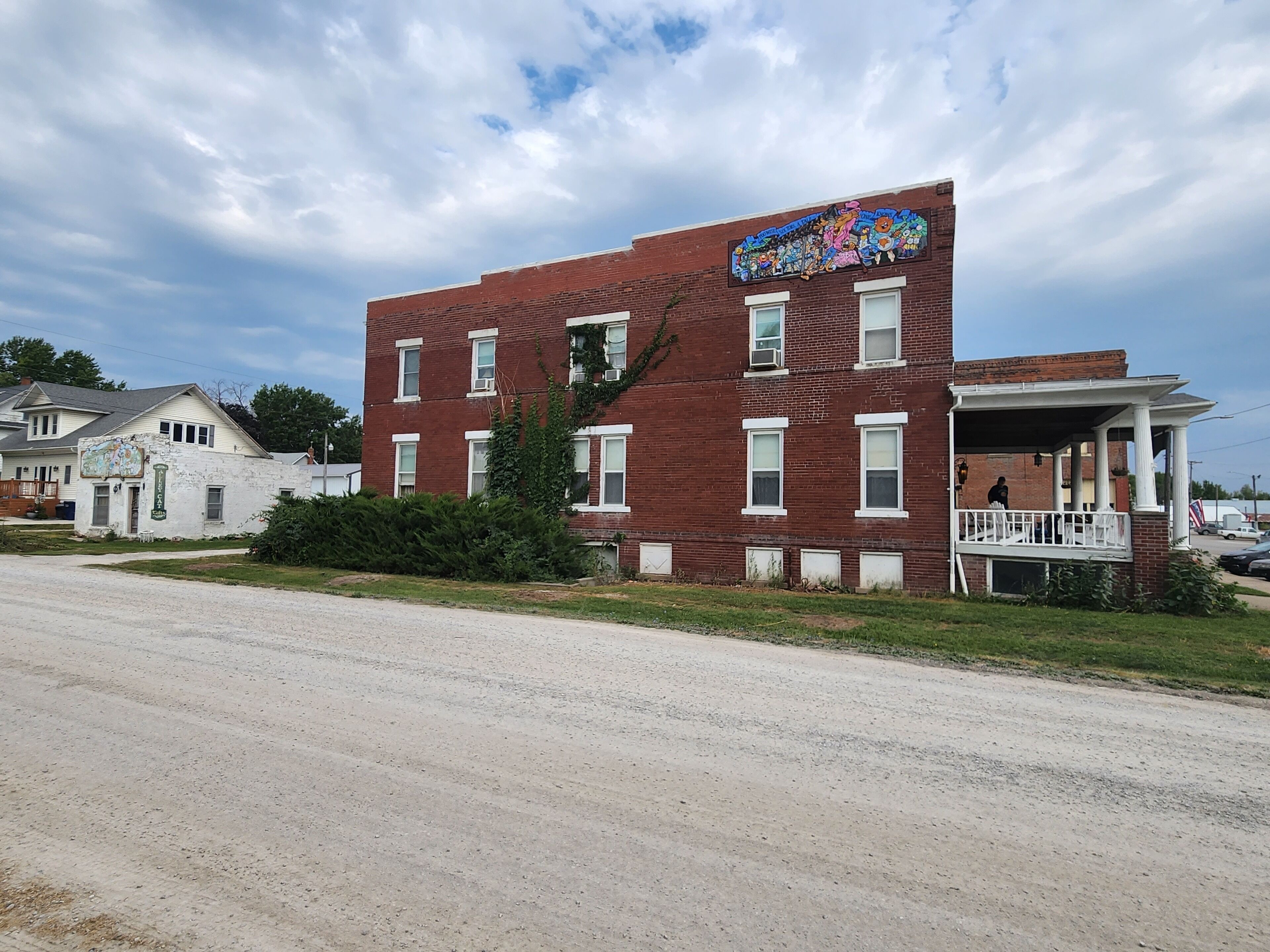 A two-story red brick building with a colorful mural on the side, a porch with white railings, and a grassy area in front.