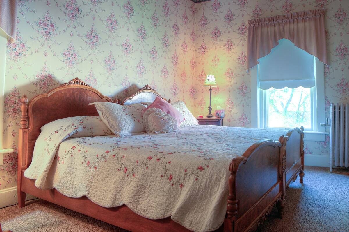 A bedroom with a wooden four-poster bed, floral wallpaper, a nightstand with a lamp, and a window with a view of trees.
