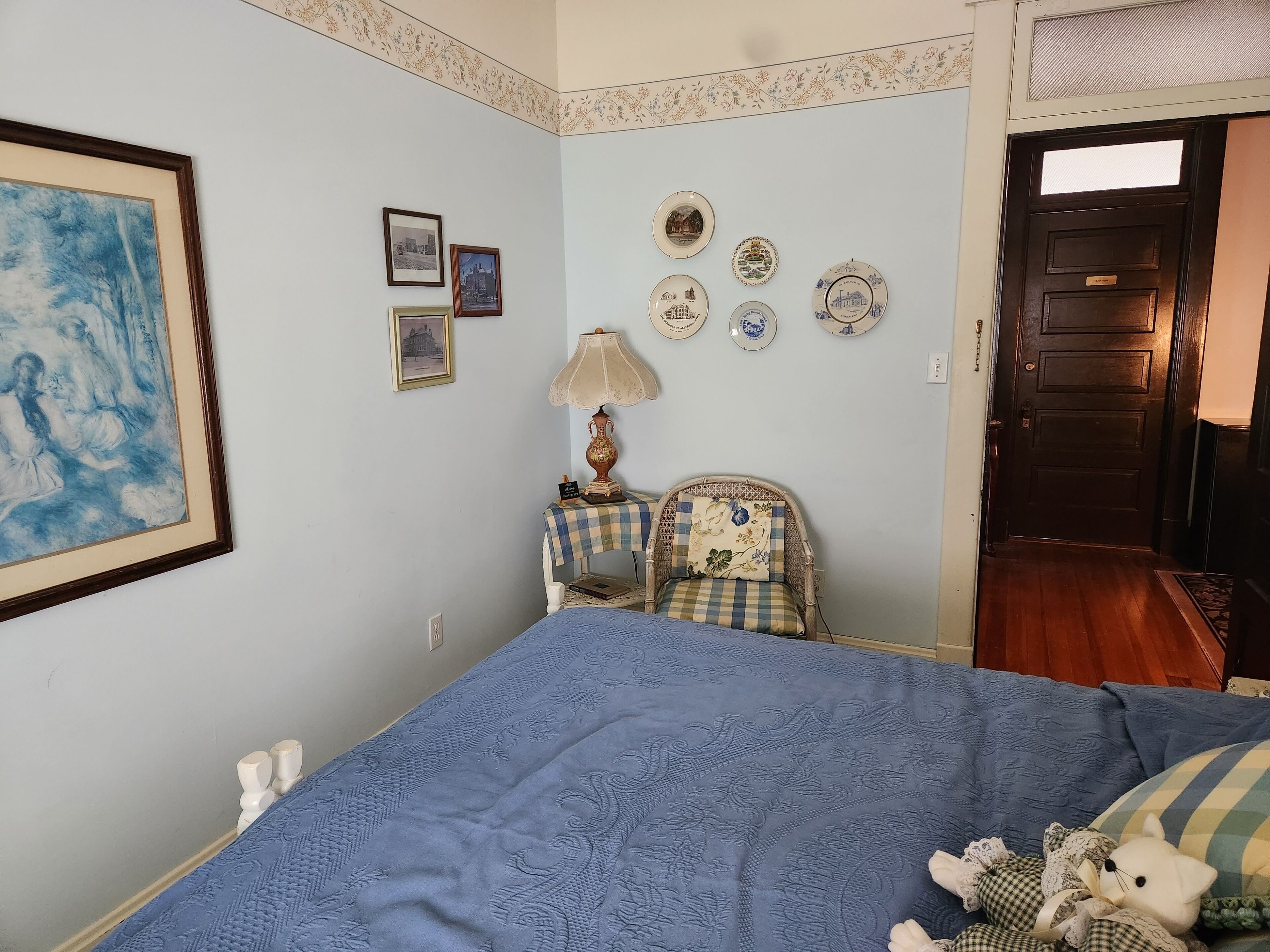 A bedroom with a bed, a chair, a lamp, and wall decorations including framed pictures and plates.