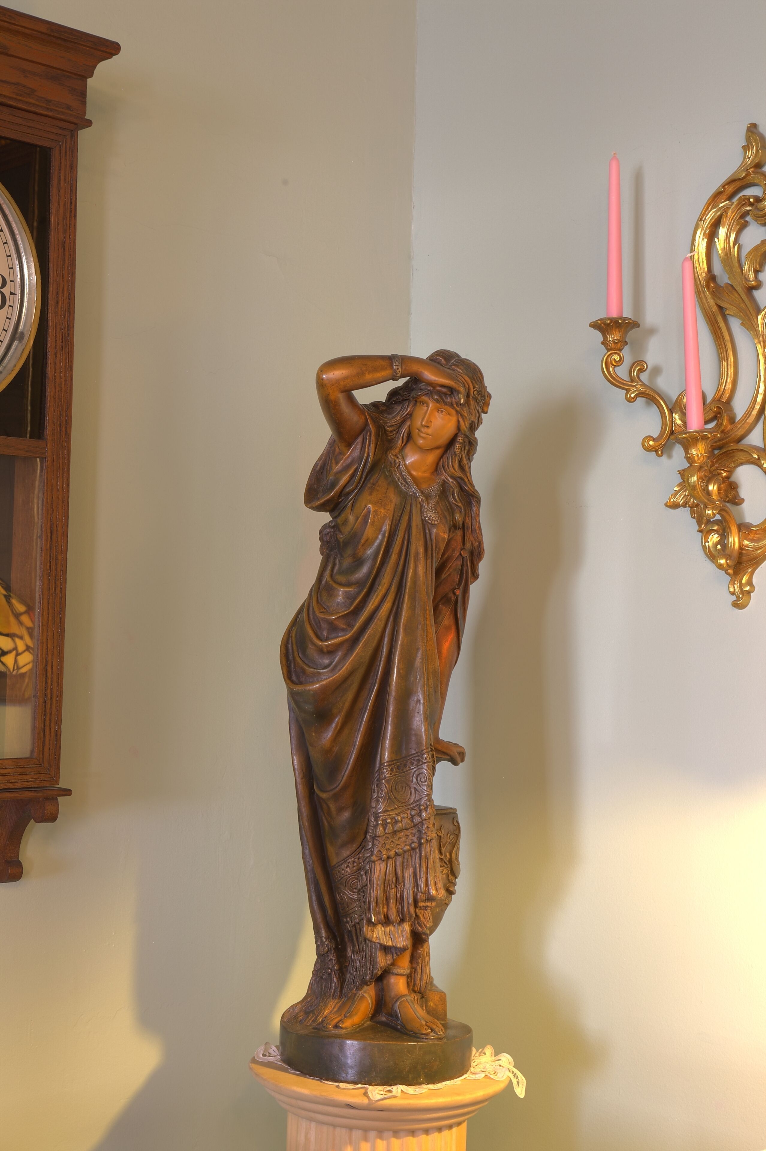 Una estatua de una mujer sobre un pedestal en una habitación con un reloj y un candelabro fijado a la pared.
