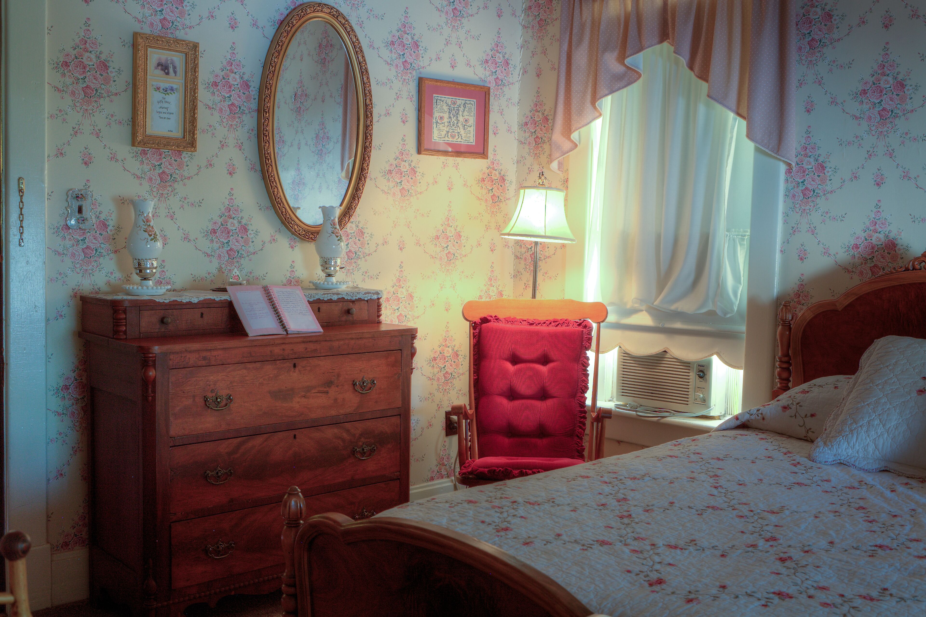 A bedroom with floral wallpaper, a wooden dresser, a red chair, a bed with a floral bedspread, a mirror, and a window with curtains.