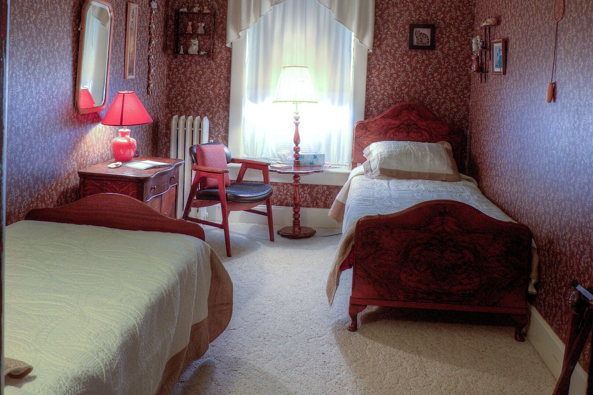 A room with two beds, a desk with a chair, a mirror, and a window with curtains.