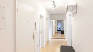 Un pasillo con paredes blancas, suelo de madera y un banco negro. Puertas alinean un lado, y un espejo refleja una habitación a lo lejos.