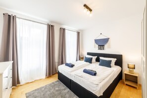 A modern bedroom with a large bed, two bedside tables, a hanging light fixture, and a view of the outdoors through sheer curtains.