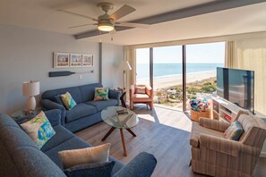 Ein modernes Wohnzimmer mit einer Couch, Sesseln, einem Couchtisch und einem Fernseher. Ein großes Fenster bietet Blick auf den Strand.