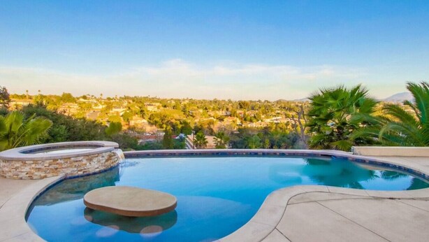 A swimming pool with a waterfall feature, surrounded by a stone deck and palm trees, overlooking a residential area.