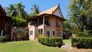 Una casa de dos pisos con techo inclinado, rodeada de abundante follaje y palmeras.