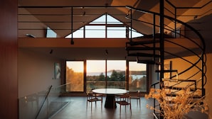 Un interior moderno con una escalera de caracol, ventanas amplias que ofrecen vistas al exterior y una mesa redonda con sillas.