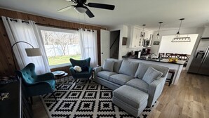 Una sala de estar moderna con sofá, sillones, un tapete estampado y una zona de cocina con armarios y un refrigerador.