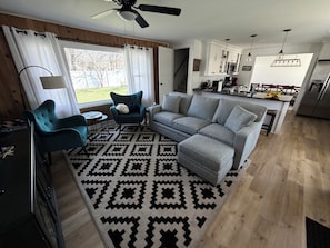 A modern living room with a sofa, armchairs, a patterned rug, and a kitchen area with cabinets and a refrigerator. - Comfortable, newly renovated home, walk to beach!
 (Michigan City)