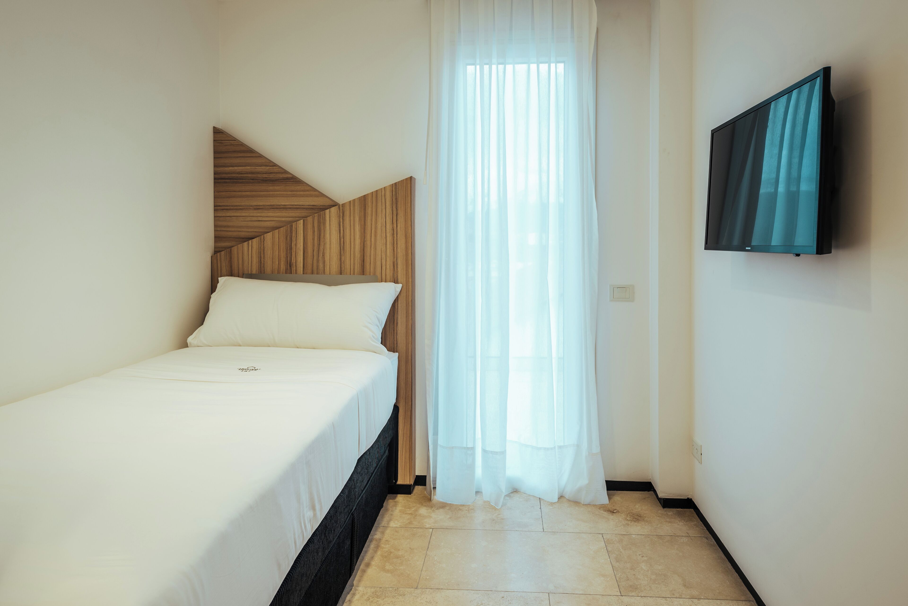 Ein kleines, ordentlich eingerichtetes Schlafzimmer mit einem Einzelbett, einem an der Wand montierten Fernseher und einem Fenster mit transparenten Vorhängen.