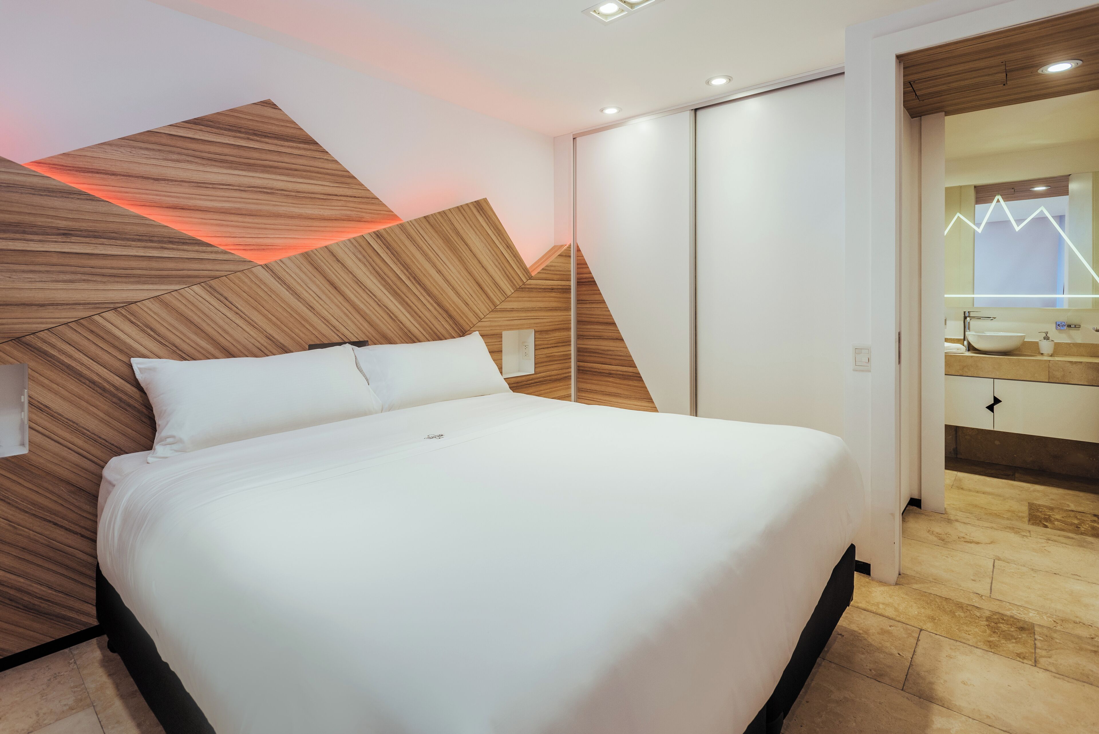 A modern hotel room with a large bed, wooden headboard, and a bathroom with a vanity and mirror.