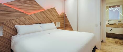 A modern hotel room with a large bed, wooden headboard, and a bathroom with a vanity and mirror.