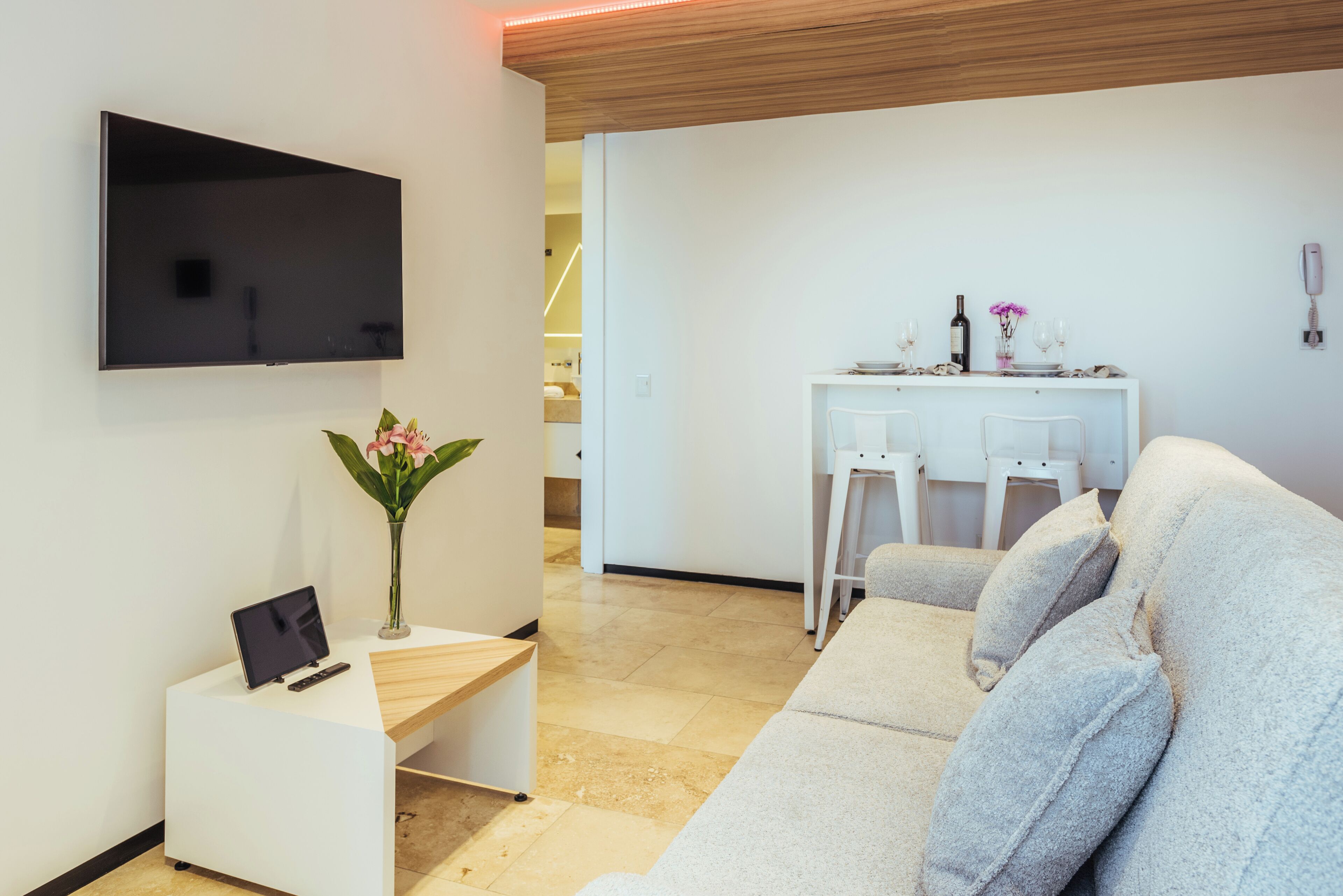 A modern living room with a flat-screen TV, a small desk with a laptop, a comfortable grey sofa, and a dining area with a white table and chairs.
