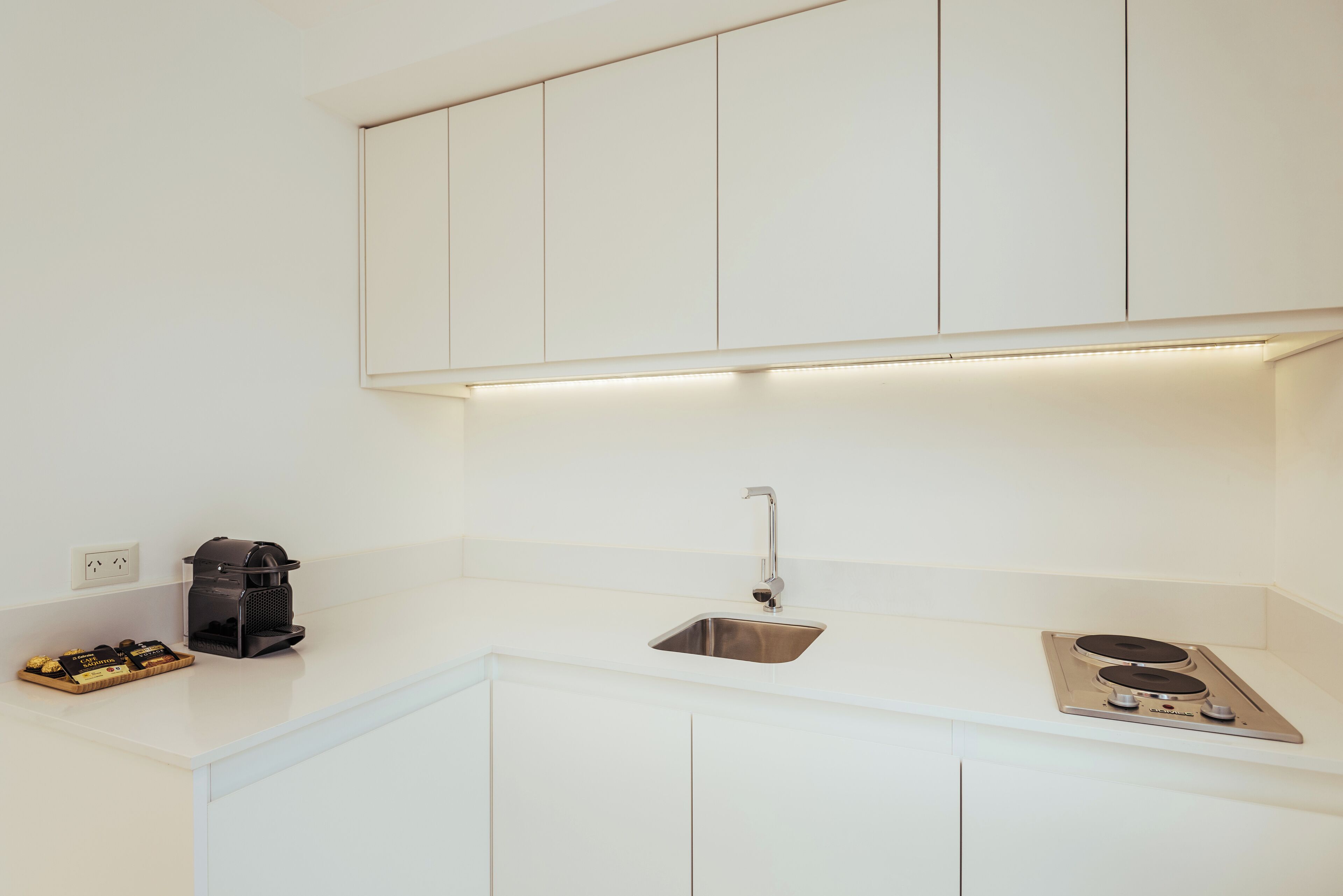 A modern kitchen with white cabinets, a stainless steel sink, a coffee machine, and a built-in stove.
