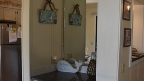 Una cocina con armarios blancos, encimera de granito y refrigerador. Hay letreros decorativos en la pared y un espejo sobre la encimera.